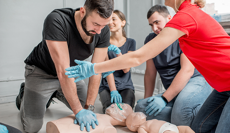 Bei einem Erste-Hilfe-Kurs beugt sich ein junger Mann über eine Übungspuppe und führt eine Herzdruckmassage durch. Neben ihm knien drei weitere Personen, eine von ihnen leitet die Übung an.