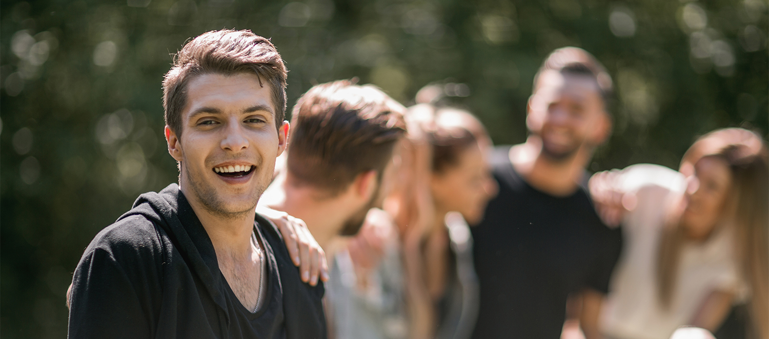 Junger Mann lacht in die Kamera. Auf seiner Schulter liegt die Hand eines weiteren jungen Mannes, der zu einer Gruppe Jugendlicher im Hintergrund des Bildes gehört.