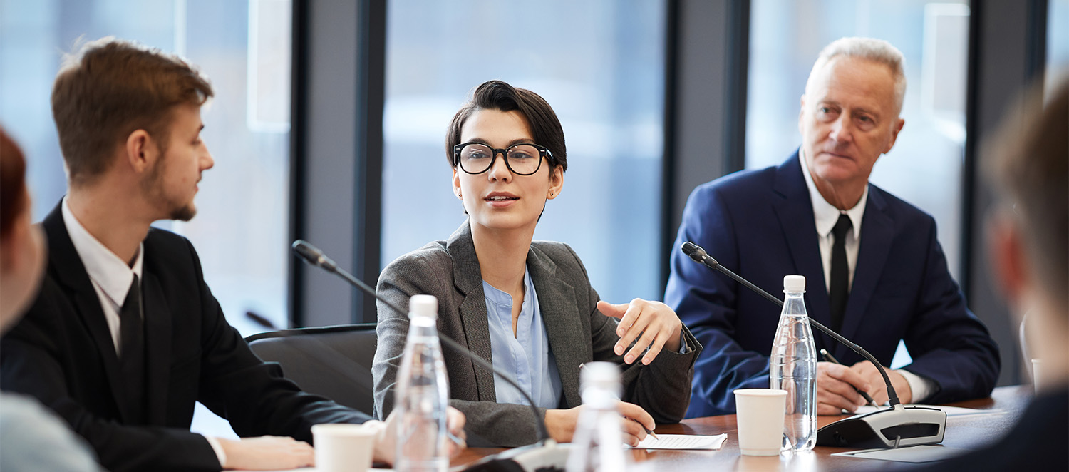 Eine Gruppe von Menschen sitzt an einem Konferenztisch mit Mikrofonen. Im Zentrum sitzt eine junge Frau, die spricht. Die zwei Männer links und rechts neben ihr hören ihr zu.