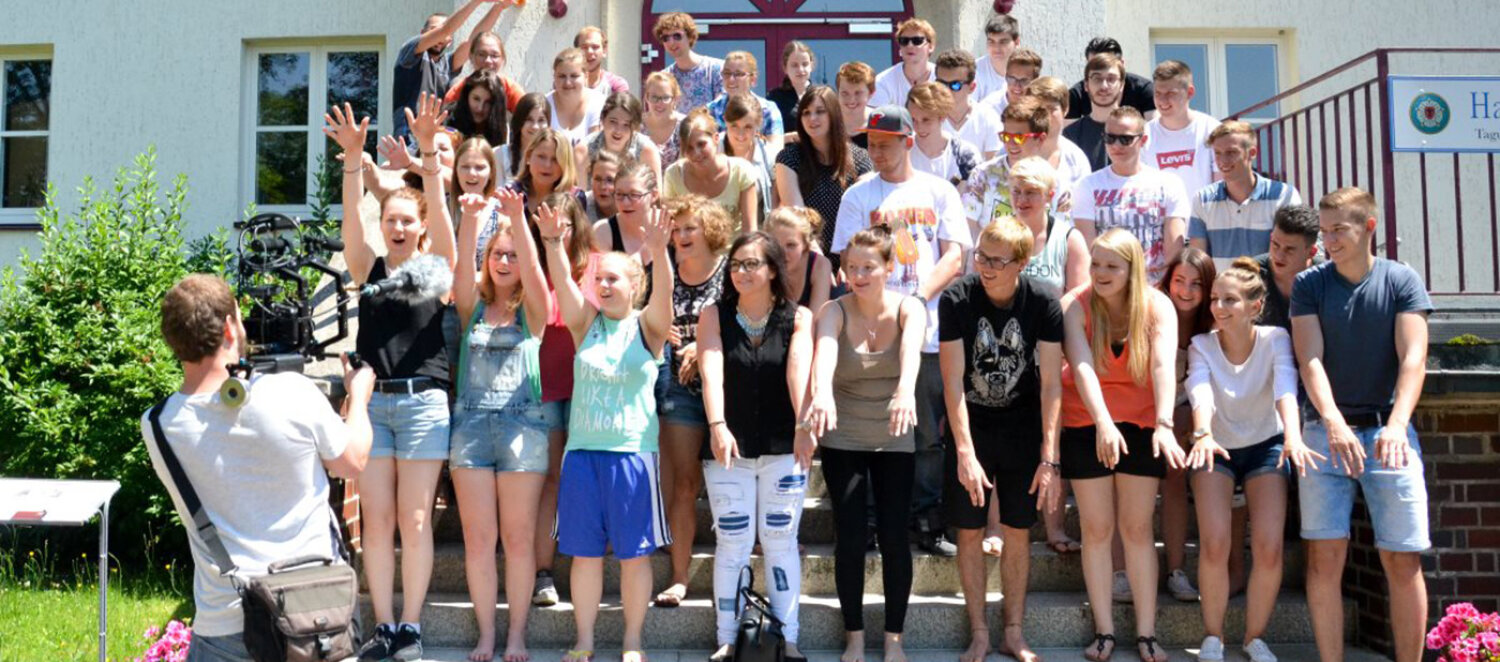 Eine Gruppe von Jugendlichen auf einer Treppe vor einem Gebäude wird von einem Kameramann gefilmt. Sie lachen und machen eine La-Ola-Welle.