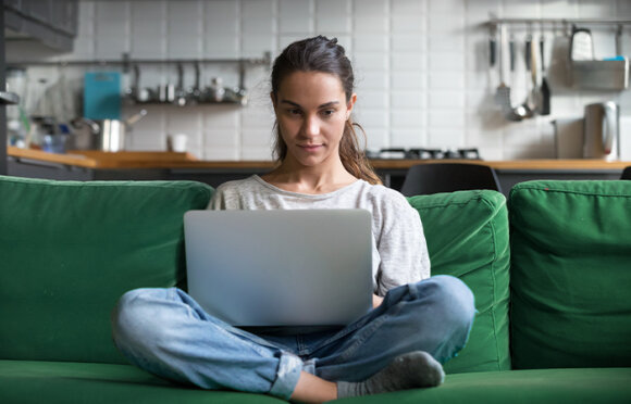 Frau sitzt im Schneidersitz auf einem grünen Sofa. Auf ihrem Schoß liegt ein geöffneter Laptop.