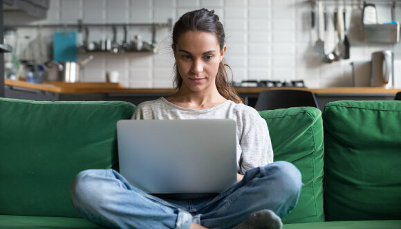Eine junge Frau sitzt im Schneidersitz auf einem Sofa. Auf ihrem Schoß liegt ein geöffneter Laptop.“