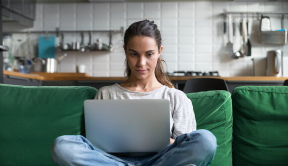 Eine junge Frau sitzt im Schneidersitz auf einem Sofa. Auf ihrem Schoß liegt ein geöffneter Laptop.