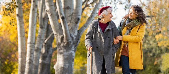 Eine junge Frau geht neben einer älteren Dame mit Gehstock durch einen Park mit Bäumen. Sie hält die ältere Frau am Arm. Beide schauen sich lächelnd an.  