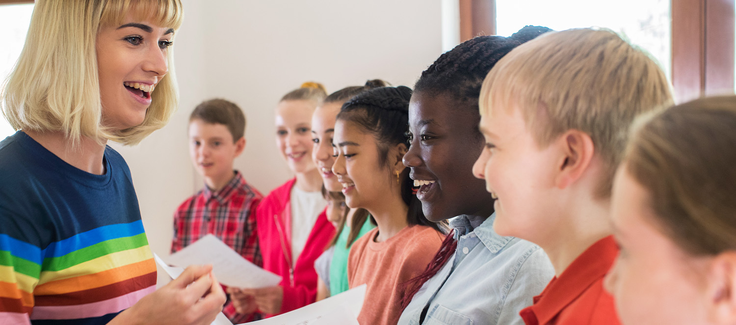 Eine junge Frau steht vor einem Chor junger Schülerinnen und Schüler, die Notenblätter in den Händen halten und singen.