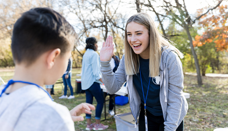 Eine junge Frau steht auf einer Wiese und begrüßt lachend einen Jungen. Sie hebt die Hand, um mit ihm abzuklatschen.