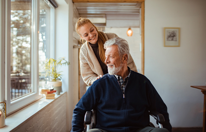 Junge lächelnde Frau steht in einer Wohnung hinter einem älteren lächelnden Mann im Rollstuhl und legt ihre Hand auf seine Schulter.