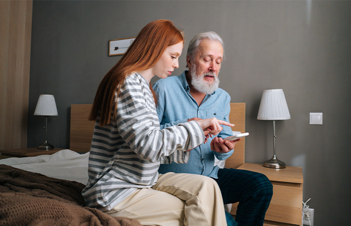 Eine junge Frau hilft einem älteren Mann bei der Bedienung seines Smartphones. Die beiden sitzen auf einem Bett.