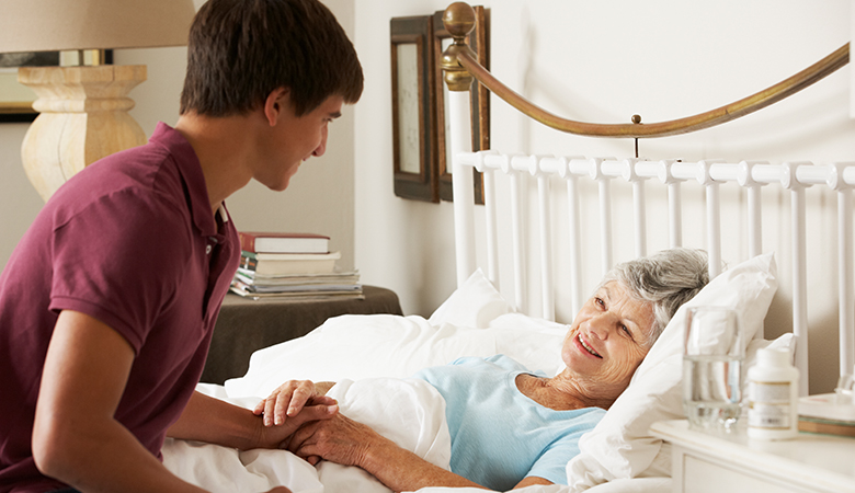 Ein junger Mann sitzt am Pflegebett einer älteren Frau. Sie halten sich an den Händen und lächeln sich an.