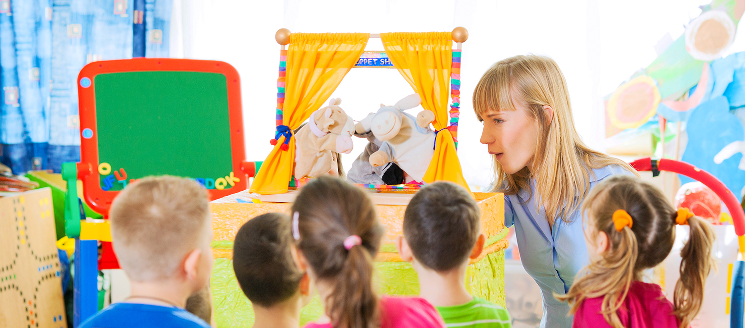 Junge Erzieherin spielt Puppentheater in einem Kindergarten vor einer Gruppe von Kindern. 