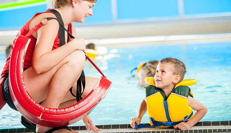 Eine junge Frau mit Rettungsring über der Schulter kniet am Rand eines Schwimmbeckens. Ein Junge mit Schwimmweste stützt sich am Beckenrand ab und schaut zu ihr auf.
