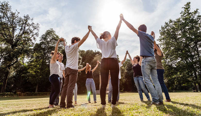 Eine Gruppe junger Menschen steht im Kreis auf einer Wiese. Sie strecken die Arme in Richtung Himmel und halten sich dabei an den Händen.