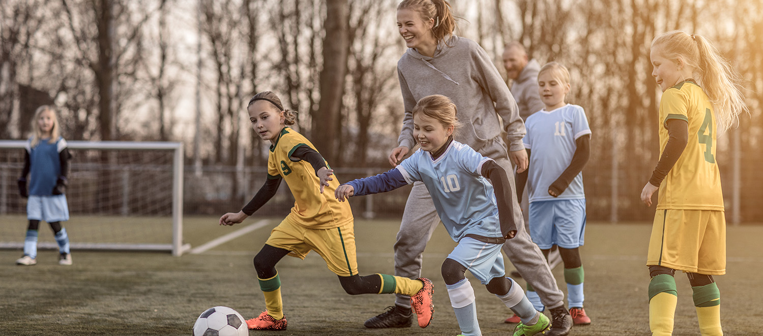 Eine junge Frau in Trainingskleidung spielt mit einer Gruppe Mädchen in Trikots Fußball auf einem Fußballplatz.