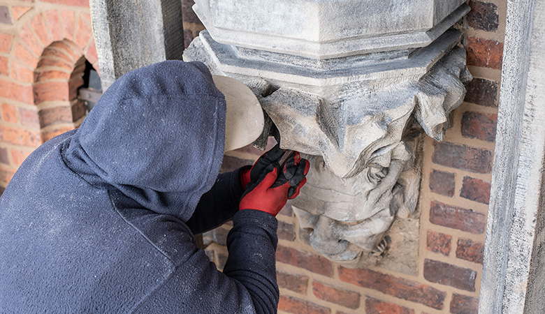 Eine Person in Arbeitskleidung arbeitet an einer Steinskulptur, die sich an einer Hausfassade befindet.