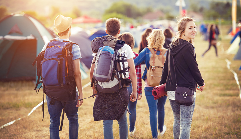 Junge Menschen mit Gepäck laufen über eine Wiese mit Zelten, die Gruppe ist von hinten zu sehen. Eine junge Frau dreht sich in Richtung Kamera und lächelt.